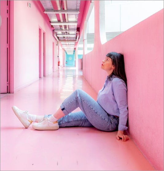 Une femme assise de profil contre un mur dans un couloir à la peinture rose